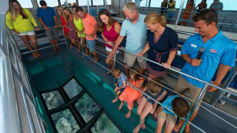 View from inside a Fury catamaran glass bottom boat showing adults and a Fury guide standing and looking down at the glass-bottom, four kids sitting with their feet dangling and also looking at the glass-bottom where they can see the ocean reef, and in the background, more adults standing and looking down at the other glass-bottom.