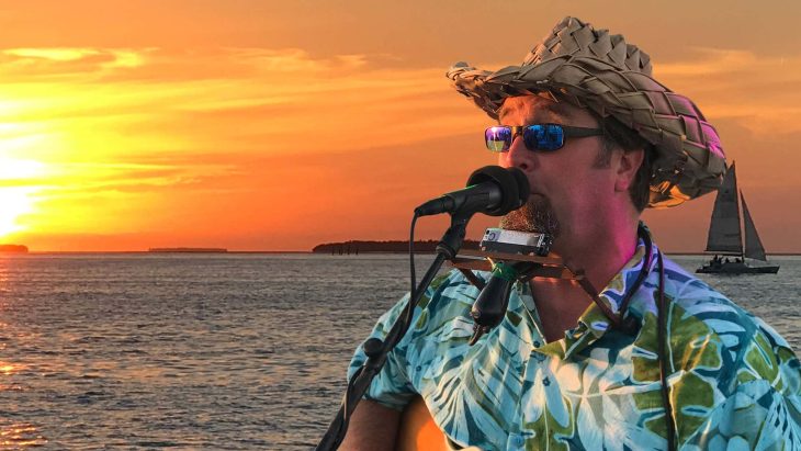 Cory Heydon wearing a straw hat and sunglasses, standing in front of a microphone and wearing a harmonica with a device holding it that wraps around his neck. He is performing Live On Fury Key West Sunset Cruise with the ocean and sunset behind him. There is also a sail boat behind him.