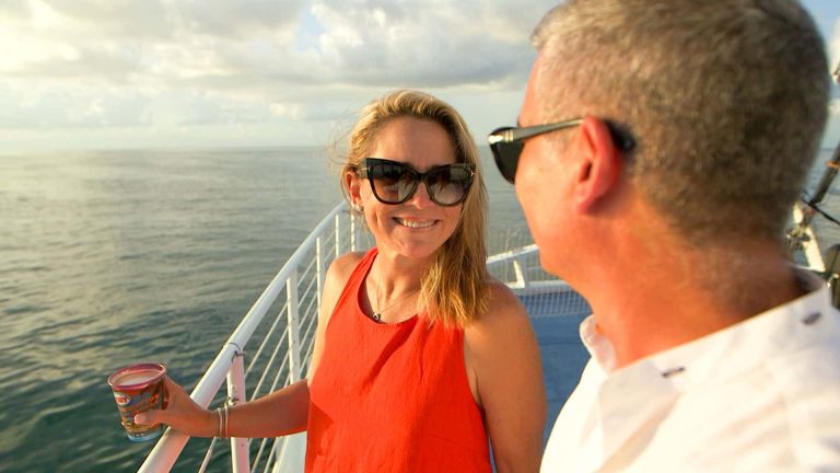Couple wearing sun glasses and looking at each other aboard Fury's Commotion on the Ocean Sunset Cruise. Both are standing next to the railing. Woman is holding a drink. And all around them is the ocean.